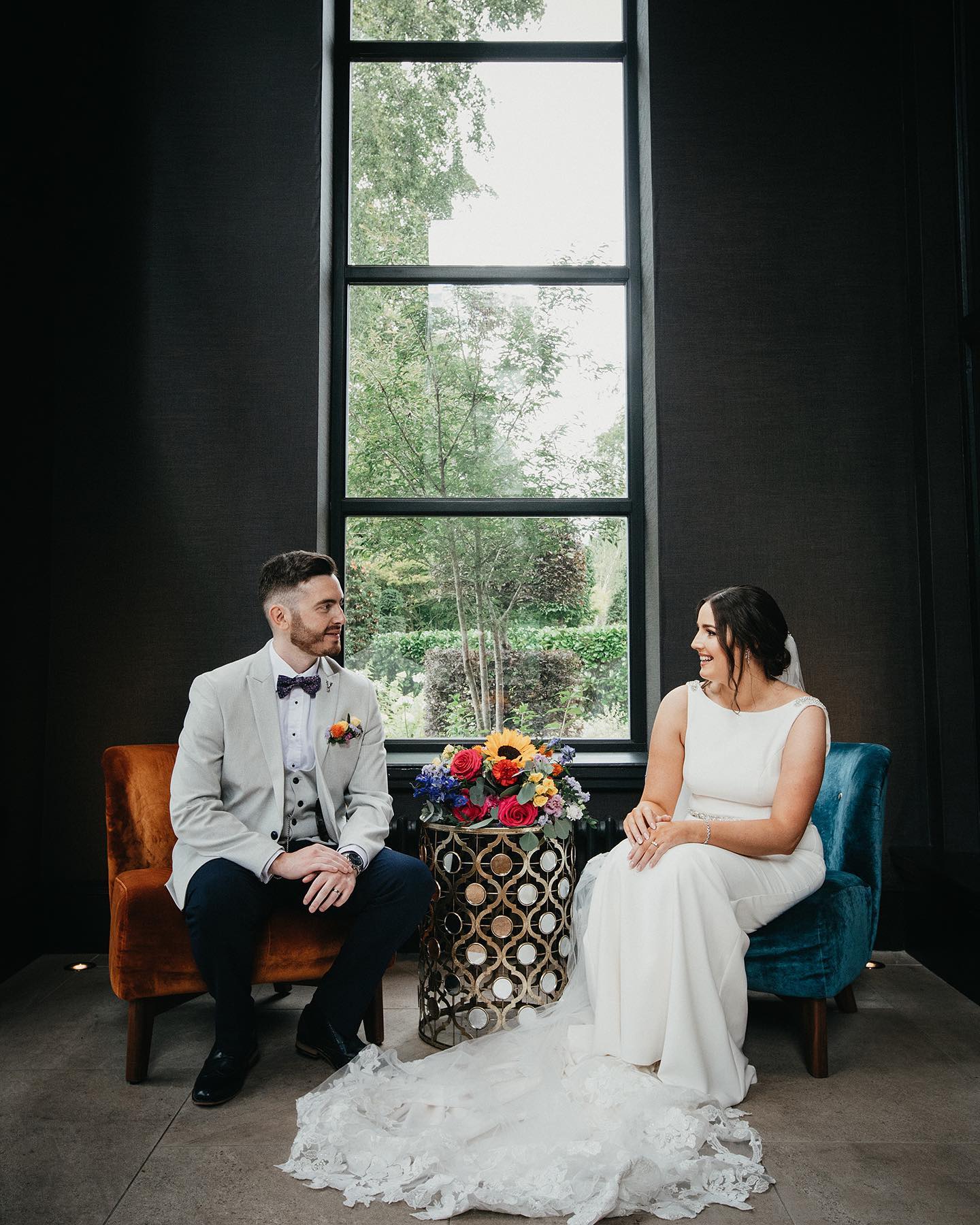 bride and groom sitting in front of a large window. A bridal bouquet on is on a table between them